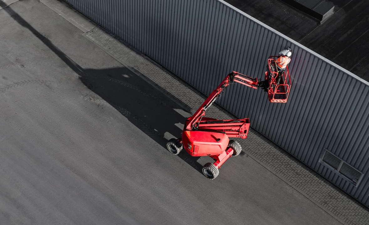 Red scissor lift with a safety-clad worker performing warehouse maintenance along an industrial loading bay, showing elevated access equipment and safe working conditions.