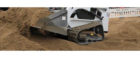 Close-up of a compact tracked loader (or a skid-steer with tracks) pushing a large pile of brown dirt and small gravel with its front bucket. The white body and black rubber track of the machine are visible