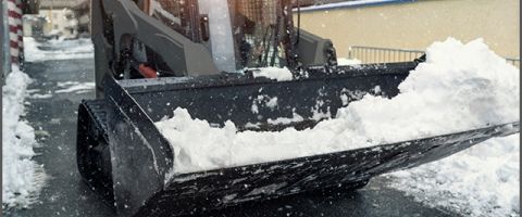 A gray compact tracked loader with snow in its bucket operates in a snowy setting